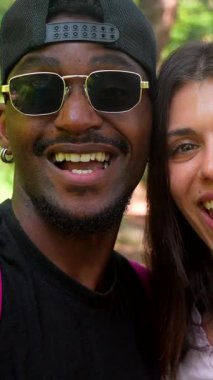 Çok ırklı genç çift, neşeli yürüyüş anlarını paylaşıyor. Güneşli orman yolunda yürürken mutluluğu yayıyorlar. Gülerek ve doğayla iletişim kurarak.