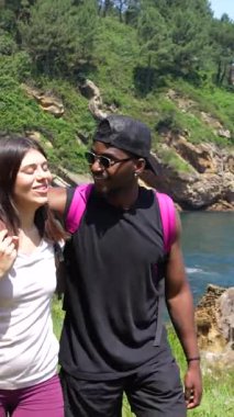 Genç çok ırklı bir çift deniz kenarında yürüyüş yapıyor, kahkahalarını paylaşıyor ve manzarayı işaret ediyor