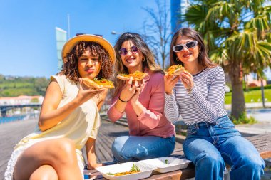 Güneşli bir parktaki bankta oturmuş pizza dilimlerinden zevk alan üç kadın kahkahalarını paylaşıyor ve keyifli bir yaz gününün tadını çıkarıyor.