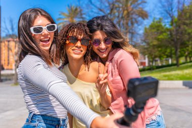Üç neşeli genç kadın barış işareti yapıyor ve şehirde yaz tatili boyunca aksiyon kamerasıyla selfie çekiyorlar.
