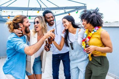 Neşeli çoklu etnik arkadaşlar çatıdaki bir partide içki içip arkadaşlıklarını kadeh kaldırarak ve kahkahalarla kutluyorlar.