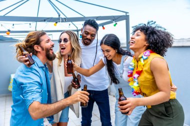 Neşeli çoklu etnik arkadaşlar bira şişeleriyle kadeh kaldırıyor, çatıdaki bir partide gülüyor ve eğleniyorlar.