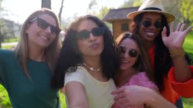 Dört çok ırklı bayan arkadaş güneşli bir parkta selfie çekiyorlar, neşe ve beraberliği ifade ediyorlar.