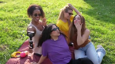 Bayan arkadaşlar yaz gününün tadını çıkarıyorlar, çimen battaniyesinde oturmuş neşeli bir selfie çekiyorlar güneşli park ortamında.