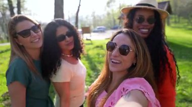 Dört çok ırklı bayan arkadaş parkta eğleniyor, grup fotoğrafları çekiyor ve gülüyorlar.