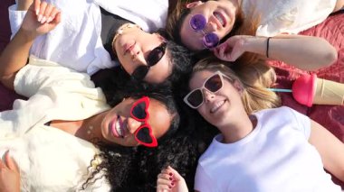 Üç neşeli genç kadın battaniyeye uzanmış güneş gözlüğü takıyor ve yaz pikniğinin tadını çıkarıyorlar.