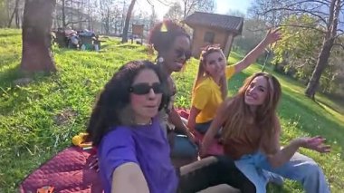Kadın arkadaşlar güneşli park pikniği sırasında neşeli anları paylaşıyorlar, yeşil çimenlerde dinlenirken gizli selfie çekiyorlar ve birlikte gülüyorlar.
