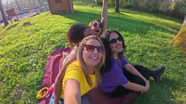 Çoklu etnik gruptan bir grup arkadaş piknik battaniyesinde oturup, yemyeşil park ortamında neşeli selfie çekiyorlar.