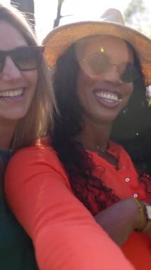 Çok kültürlü bayan arkadaşlar açık hava eğlencesi anlarında kaynaşıyor, lüks park yeşillikleriyle çevrili moda günlük giysiler giyerken neşeli selfie çekiyorlar.