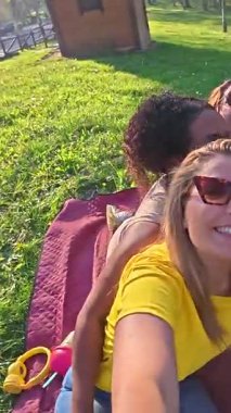 Bayan arkadaşlar dışarıda toplanıyor, battaniyeye oturup selfie çekiyorlar, birlikte gülüyorlar ve yemyeşil parkta güneşli bir günün tadını çıkarıyorlar gerçek mutluluk ve kaygısız ruhla.