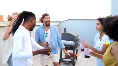 Çok ırklı arkadaşlar yaz akşamını çatıda kutluyorlar, enerjik bir şekilde dans ediyorlar, içkilerini ve kahkahalarını paylaşıyorlar şehrin ufuk çizgisinin ve şenlikli atmosferinin tadını çıkarıyorlar.