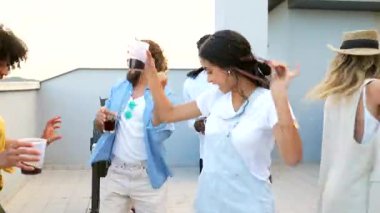 Çok kültürlü arkadaşlar çatıdaki toplantı sırasında canlı anılar yaratıyor, kahkahalar paylaşıyor, dans ediyor, şehir ışıkları altında kokteyllerin tadını çıkarıyor.