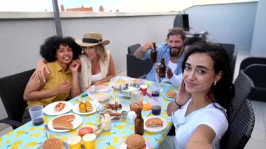 Çok kültürlü genç arkadaşlar barbeküyü paylaşırken hatırlanmaya değer anlar yaratıyor, canlı çatı katındaki sosyal ortamlarda selfie çekiyorlar.