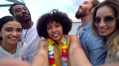 Neşeli çoklu etnik arkadaşlar çatıdaki bir partide renkli kıyafetler ve çiçek kolyeleriyle selfie çekiyorlar.