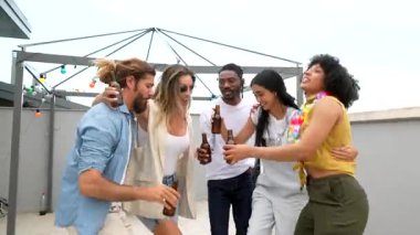 Çok ırklı genç arkadaşlar yaz akşamını dans ederek, kadeh kaldırarak ve şehir terasında gece ışıkları altında içki içerek kutluyorlar.