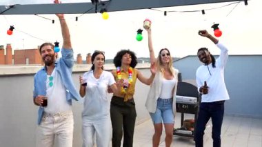 Enerji dolu çoklu etnik arkadaşlar dans ediyor, içkilerini paylaşıyor ve yaz akşamları canlı çatı partisi atmosferinin tadını çıkarıyor.