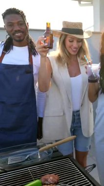 Canlı bir grup arkadaş çatıdaki barbeküden zevk alıyor, lezzetli yemekler pişiriyor ve altın gün batımı ışığı altında içeceklerle kadeh kaldırıyor.