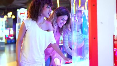 Yaz eğlencesi, genç kadınların atari oyunuyla bağ kurması, renkli oyun ortamının tadını çıkarırken kahkaha ve heyecanı paylaşması.