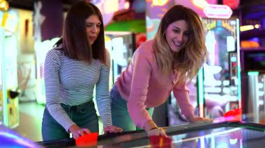 Dost canlısı hava hokeyi maçına katılan genç kadınlar, renkli atari salonu içinde yaz neşesi ve enerjik eğlence yayıyorlar.