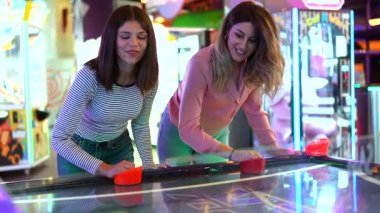 İki genç kadın hava hokeyi oynarken yarışıyor ve gülüyorlar. Etrafları renkli atari ışıkları ve canlı eğlence enerjisi yakalıyorlar.