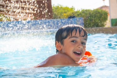 Gülümseyen çocuk, ferahlatıcı bir şelalenin altında şişme kolluklarıyla mavi bir yüzme havuzunda oynayarak yaz tatilinin tadını çıkarıyor.