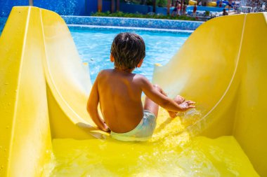 Küçük çocuk yaz tatilinin tadını çıkarıyor. Sarı su kaydırağından su parkındaki yüzme havuzuna kayıyor.