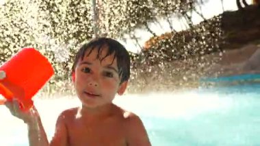 Parlak turuncu kovayla neşe içinde su sıçratan genç çocuk güneşli gökyüzünün altında serinletici havuz suyunun keyfini çıkarırken keyifli yaz anları yaşıyor.