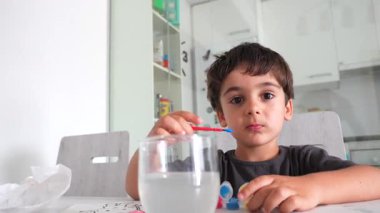 Sanatsal beceriler geliştiren genç bir çocuk, parlak mutfakta beyaz masalarda suluboya resimler yapıyor, oyuncu öğrenme anında konsantrasyon ve yaratıcılık sergiliyor.