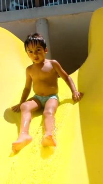 Enerjik genç çocuk canlı sarı su kaydırağından kayıyor, güneşli bir yaz günü su parkında serin mavi havuza neşeyle sıçrıyor.