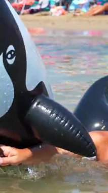 Neşeli çocuk yaz tatili boyunca kumlu sahilde parlak güneş ışığında yüzerken gülüyor ve şişme balinalara sarılıyor.