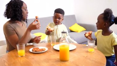 Afrikalı Amerikalı anne çocuklarıyla kahvaltı yapıyor, sıcak ve sevgi dolu bir atmosferde yemek ve kahkaha paylaşıyor.