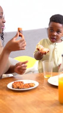 Afro-Amerikalı genç bir çocuk besleyici bir kahvaltıyı şefkatli bir anneyle paylaşıyor, sağlıklı yemeğin tadını çıkarırken aynı zamanda mutfakta sıcak bir aile bağı oluşturuyor.