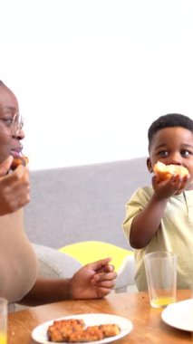 Çocuk mutlu bir şekilde aile masasında sandviçini yiyor, yemek zamanını anne ile mutfak ortamında paylaşıyor.