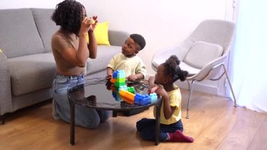 Anne, oğlu ve kızıyla etkileşime girer. Sıcak bir oturma odasında renkli yapı taşlarıyla oynar.