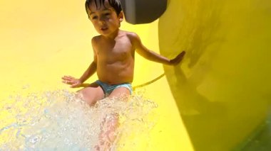 Parlak sarı su kaydırağında kayan genç çocuk, tatil köyünde neşeli yüzme anları yaşıyor neşeli bir heyecan ve kaygısız enerji ile