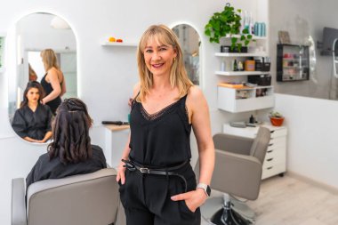 Kendine güvenen kadın girişimci elleri ceplerinde modern kuaför salonunu yönetirken müşteri yeni bir saç stili alıyor.