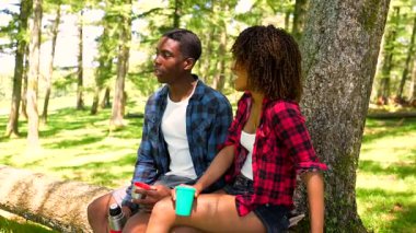 Genç yürüyüşçüler ormanda mola veriyor, düşmüş bir ağaç gövdesine oturuyor, içki içiyor ve sohbet ediyorlar.