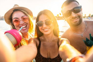 Bir grup neşeli çoklu etnik arkadaş plaj partisinde selfie çekiyor, gün batımında taze içkilerin tadını çıkarıyorlar.