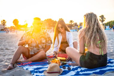 Gün batımı pikniğinde plaj havlusunda karpuz ve ananasın tadını çıkaran bir grup arkadaş, canlı bir yaz sahnesi yaratıyor.