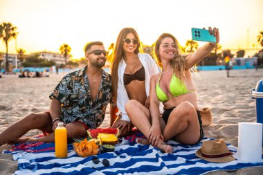 Üç arkadaş gün batımında plaj pikniğinde taze meyve ve atıştırmalıklarla selfie çekiyorlar.