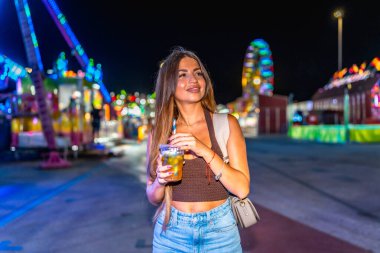 Genç bir kadın neşeli bir lunaparkta içkisini yudumluyor, renkli ışıklarla aydınlatılıyor, neşeli bir ortam yaratıyor.