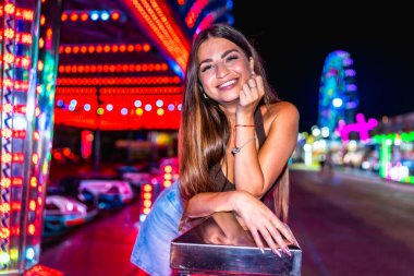 Eğlence parkında eğlenen mutlu bir kadın. Renkli çarpışan arabalarla tırabzanlara yaslanıyor ve arka planda dönme dolap var.