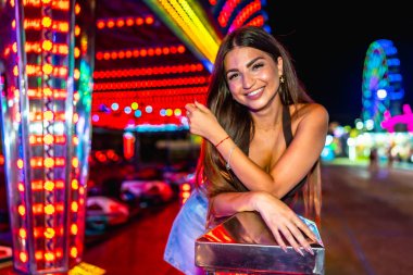 Bir lunaparkın canlı atmosferinin tadını çıkaran mutlu genç bir kadın, geceleri renkli bir şekilde poz veriyor.