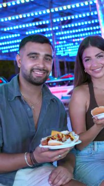 Gece karnavalı şenliklerinde genç arkadaşlar sosisliden zevk alıyor, renkli ışıklandırılmış park ışıkları altında kahkahalar atıyorlar.