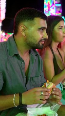 Genç arkadaşlar, canlı bir eğlence parkında sosisli ve hamburger yiyerek, canlı atmosferin tadını çıkarıyorlar.