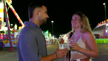 Gece vakti eğlence parkı sahnesinde genç bir çift gülüyor, içkilerini paylaşıyor renkli ışıklar ve şenlikli enerjiyle canlı atmosferin tadını çıkarıyor.