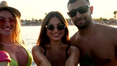 Üç arkadaş, iki kadın ve bir erkek sahilde güzel bir gün batımında birlikte kokteyl içerler.