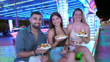 Parlak bir lunaparkta sosisli yiyen mutlu arkadaşlar, birlikte eğleniyorlar.
