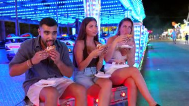 Arkadaşlar lezzetli sosisleri paylaşıyor, birlikte gülüyor ve gece vakti eğlence parkı deneyimi sırasında karnaval eğlencesine katılıyorlar.