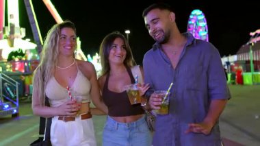 Genç yetişkinler gece canlı bir eğlence parkında gezerler, ferahlatıcı içeceklerin tadını çıkarırlar ve canlı sohbetlerle meşgul olurlar.